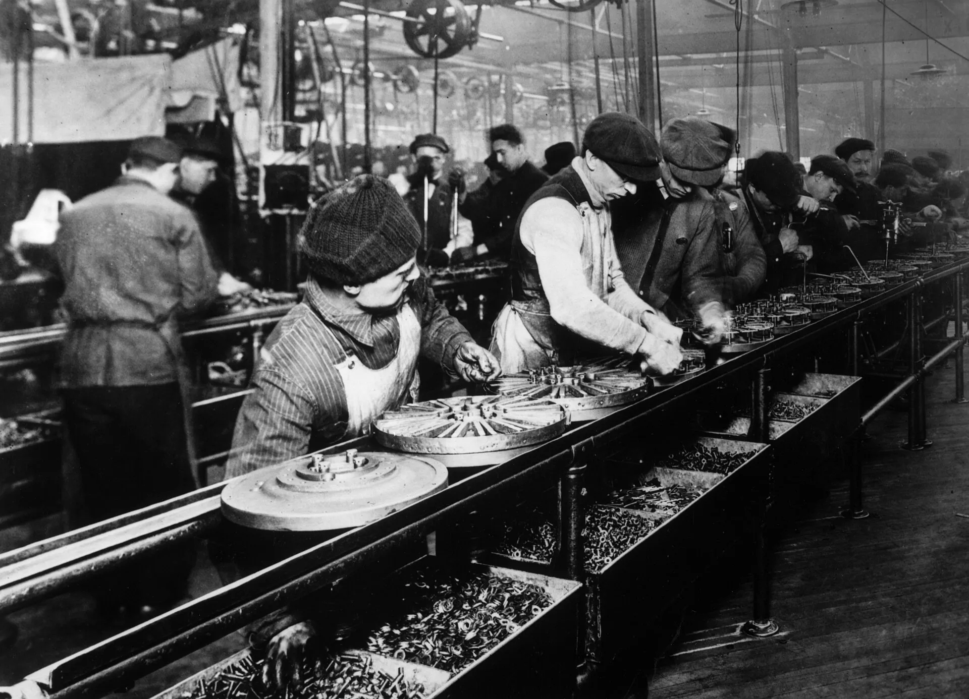 Workers in caps stand along a long assembly line in a Ford factory in 1913, each repeating one operation on parts that move past them.